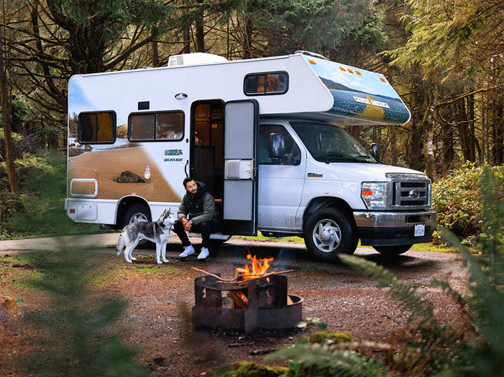 Man sitting in door of RV with husky dog near a fire pit