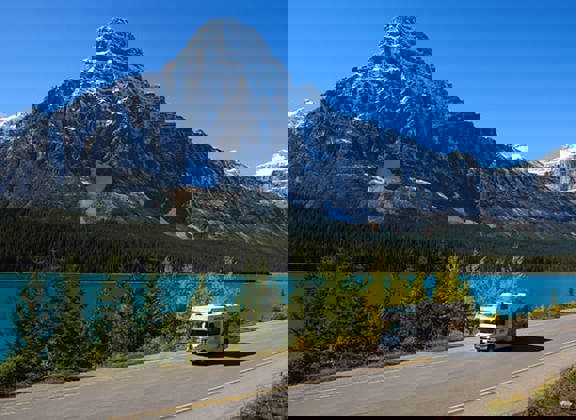 RV on road in front of blue lake and mountain.